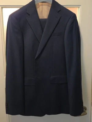 Mörkblå kostym från Bläck - Snygg mörkblå kostym från Bläck med modern skräddad passform. Kavajen har klassiska slag, två framfickor med lock och en bröstficka. Beige innerfoder och diskret märkning. Perfekt för dig som vill ha en stilren look. Kavaj: Stl 48 Byxor: Stl 44