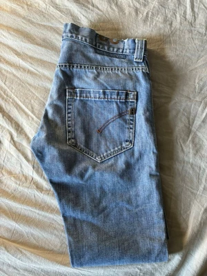 Dondup jeans - Säljer ett par ljusblå jeans med rak passform och klassisk femficksdesign. Jeansen har en bakficka med dekorativ söm och bälteshällor i midjan. Tillverkade i denim av bomull för en avslappnad look.