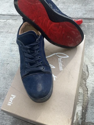 Blå sneakers från Christian Louboutin - Snygga blå sneakers från Christian Louboutin med klassisk röd sula och lågt skaft. Skorna har ovandel i mocka och detaljer i läder, samt matchande blå skosnören. Perfekta för dig som vill sticka ut med exklusiv streetstyle.