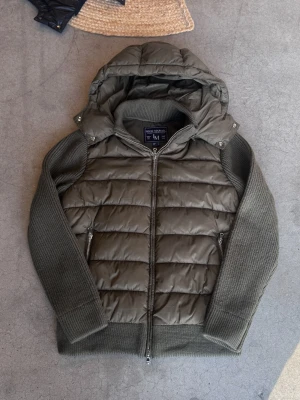 Marcél Classic Hybridjacka – Olive | Avtagbar Luva | Ny Med Prislapp - Ny med prislapp — Marcél Classic i djup grön färg med quiltad pufferkropp och stickade ärmar. Avtagbar luva och lyxiga microfleecefickor som håller händerna varma — detaljer som gör skillnad. Perfekt för både höst/vinter och kyliga vårdagar — en jacka du bär hela säsongen. Finns i flera storlekar — skriv din längd och vikt så hjälper jag dig hitta den perfekta passformen! Säljer direkt för ett bättre pris än butik. 📦 Spårad frakt, väl förpackad.