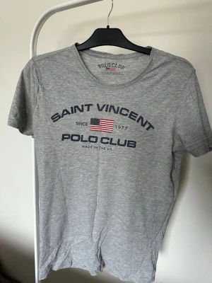 2-pack Saint Vincent Polo Club t-shirts - Två t-shirts från Saint Vincent Polo Club, en i ljusgrått och en i marinblått. Båda har rund hals, korta ärmar och tryck med amerikansk flagga och text framtill. Perfekta för en avslappnad och sportig stil.