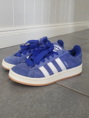 Blå Adidas Campus sneakers - Säljer ett par blå Adidas Campus sneakers med klassiska vita ränder på sidan och vit logga på tungan. Skorna har en låg profil, breda blåa skosnören och en ljus gummisula. Ovandelen är i mocka vilket ger en snygg och stilren look. 