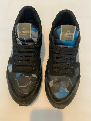 Valentino rockrunners storlek 42 - Snygga sneakers från Valentino Garavani med svart, blå och grå camomönstrad ovandel. Skorna har svarta mockadetaljer, svarta snören och coola nitar på sulan. Perfekt för dig som vill sticka ut med en unik och trendig look.                                                               Kom med bud!😃