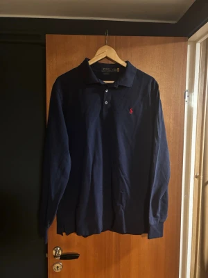 Polo Ralph Lauren Piké - Säljer en marinblå långärmad piké från Polo Ralph Lauren med klassisk krage och knappar framtill. Skjortan har det ikoniska röda Polo-logotypen broderad på bröstet och är tillverkad i mjuk bomull. Perfekt för dig som gillar stilrena plagg.