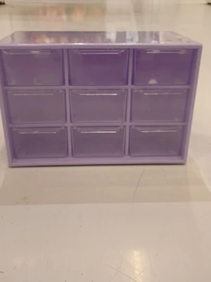 Lila förvaringslåda med 9 fack - Praktisk förvaringslåda i lila plast med nio små utdragbara fack. Perfekt för att organisera småsaker som smycken, kontorsmaterial eller hobbyprylar. Lådan har en rektangulär form och kan även hängas på väggen tack vare två hål på baksidan.