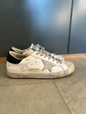 Golden Goose vita sneakers  - Golden Goose sneakers i vitt läder med grå mockastjärna på sidan och svart hälparti. Skorna har gråa skosnören och en blå detalj på plösen. Klassisk låg modell med vintage-inspirerad design och GGDB/SSTAR-tryck på sidan.