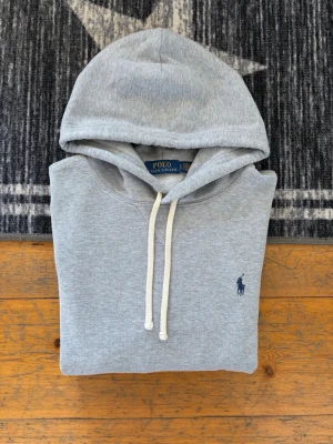 Ljusgrå Polo Ralph Lauren Hoodie - Hej! 👋 Säljer en klassisk ljusgrå (grey heather) Polo Ralph Lauren hoodie i storlek L (Herr). Den har den ikoniska mörkblå pony-loggan broderad på bröstet 🐎 Tröjan är i mycket fint skick, mjuk, skön och minimalt med noppor. Denna modell har QR-kod för enkel verifiering av äkthet directly ✅ Observera att de långa tvättlapparna är avklippta för komfort, men alla andra etiketter i nacken sitter kvar 👕 Ett tidlöst premium-basplagg som passar perfekt året om. Fraktar snabbt! 🚛📦💬💯 