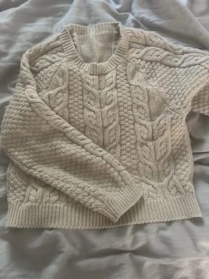 Beige kabelstickad tröja - Säljer en beige kabelstickad tröja med klassiskt flätmönster och ribbade muddar. Tröjan har rund hals och är gjord i mjukt material som passar perfekt för kyliga dagar. 