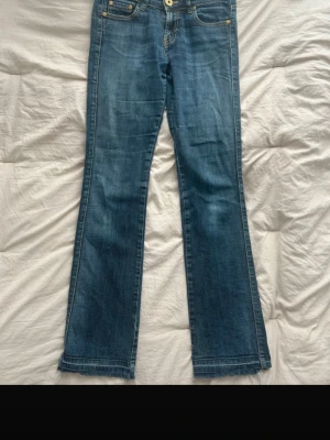 Blå bootcut jeans med lågmidja - Säljer ett par blå lågmidjade bootcut jeans med klassisk femficksdesign och rå, fransad kant nedtill. Jeansen har en snygg tvättad look och är tillverkade i denim med lätt stretch. Perfekta för en avslappnad och trendig stil. De är något slitna men sitter perfekt i midjan och i längden.