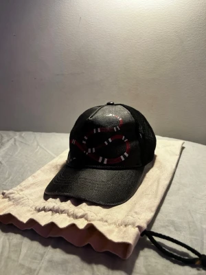 Svart keps med orm från Gucci - Cool svart keps från Gucci med mesh baktill och justerbar rem. Framtill finns en snygg broderad röd, vit och svart orm som ger kepsen en unik look. Materialet är en mix av bomull och mesh, perfekt för dig som vill sticka ut.