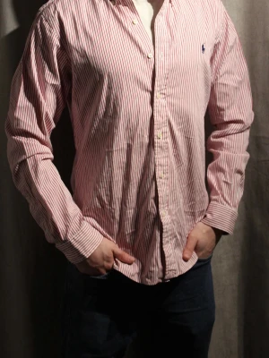 Rödvit randig skjorta från Ralph Lauren - Klassisk skjorta från Polo Ralph Lauren i rödvitt randigt mönster. Skjortan har lång ärm, knappar framtill och en liten blå logga broderad på bröstet. Tillverkad i bomull och har en normal passform. Perfekt för en clean och stilren look.