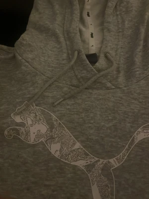 Grå hoodie från Puma med tryck - Säljer en gråmelerad hoodie från Puma med stor logga framtill. Hoodien har huva med dragsko och en klassisk magficka. Materialet är mjukt och bekvämt, perfekt för chill dagar. Passar dig som gillar sportig och avslappnad stil.