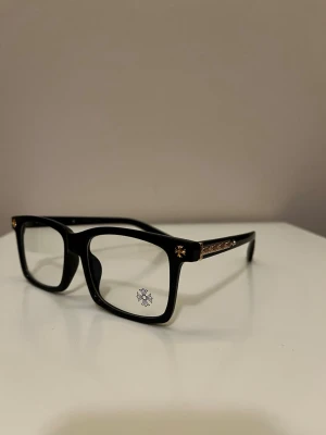 Chrome hearts Svarta blue light glassor med guldiga detaljer - Snygga solglasögon med bred svart båge och fyrkantig form. Glasögonen har guldiga dekorationsdetaljer på sidorna och vid hörnen, vilket ger en lyxig känsla. Perfekt för dig som vill sticka ut och addera lite bling till din look.