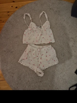 Sött vitt pyjamasset med rosa blommor - Tvådelat pyjamasset i vitt med små rosa blommor. Toppen har smala axelband med volangdetaljer och knytning fram. Shortsen har resår i midjan, knytband med hjärtformade detaljer och volangkant nedtill. Materialet ser ut att vara mjukt och luftigt, perfekt för mysiga kvällar.