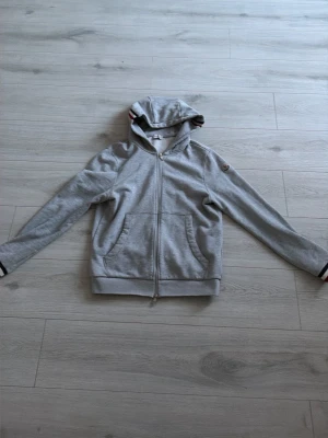 Grå zip-hoodie från Moncler - Snygg grå hoodie från Moncler med dragkedja och huva. Klassisk logotyp på ärmen och detaljer med röd och vit rand på huvan. Mjuk bomullskänsla och praktiska fickor framtill. Perfekt för en avslappnad och cool look.