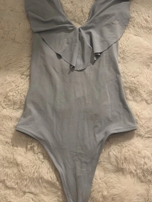 Ljusblå offshoulder body med volang - Ljusblå body med turtleneck-design och bred volang framtill. Materialet är mjukt och stretchigt, vilket ger en bekväm passform. Bodyn har en djup v-ringning och är ärmlös, perfekt för en trendig och stilren look. Ingen synlig logga eller märke.