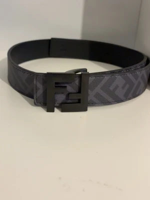 Grå Fendi bälte med FF-mönster - Snyggt grått bälte från Fendi med ikoniskt FF-mönster och en cool metallspänne med Fendi-logga. Bältet har en bred och stilren design som passar perfekt till jeans eller kostymbyxor. Materialet är skinn och detaljerna är i mörk metall.