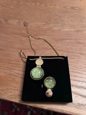Guldfärgad ring och halsband med grön detalj - Unik guldfärgad ring och halsband, ring och halsband har en glittrande grön yta som fångar ljuset snyggt. Ringen har en modern och stilren design som sticker ut och passar perfekt för dig som gillar statement-smycken.