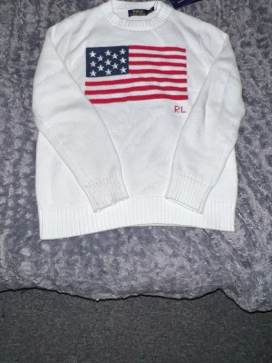 Vit stickad tröja Polo Ralph Lauren - Vit stickad tröja från Polo Ralph Lauren med amerikansk flagga i blått, vitt och rött på bröstet samt RL-broderi. Tröjan har rund hals, långa ärmar och ribbstickade muddar vid ärmslut och nederkant. Perfekt för dig som gillar klassisk stil.