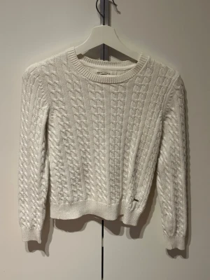 Vit kabelstickad tröja Holly & Whyte - Säljer en vit kabelstickad tröja från Holly & Whyte med rund halsringning och ribbade muddar. Tröjan har ett klassiskt flätmönster och är perfekt för att hålla sig varm under kyliga dagar. Materialet är mjukt och bekvämt.