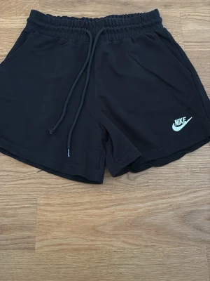 Svarta Nike shorts med snörning - Säljer ett par svarta shorts från Nike med elastisk midja och snörning. De har en klassisk logga i vitt på ena benet och är tillverkade i mjukt material som passar perfekt för träning eller chill. Modellen är loose fit och har praktiska sidofickor.