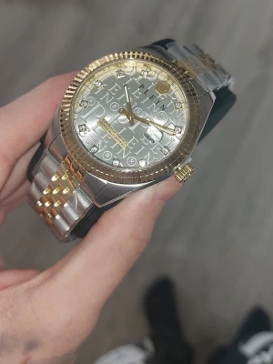 Philipp Plein silver-guld klocka - Snygg klocka från Philipp Plein med silverfärgad urtavla och guldiga detaljer. Armbandet är tvåfärgat i silver och guld, och urtavlan har ett coolt mönster med bokstäver. Perfekt för dig som vill sticka ut med en lyxig accessoar. Finns länkar så det kan justeras så den passar just dig! Väldigt fin klockan och säljer den med en tår i ögat har haft den cirka några månader ser ut som ny, kvittot finns digitalt och det önskas! Bara höra av dig för vidare information Riki!!