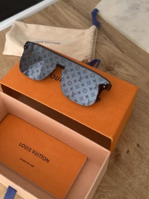 Louis Vuitton monogram solglasögon - Säljer ett par exklusiva solglasögon från Louis Vuitton med ikoniskt monogrammönster på linsen. Glasen är mörkgrå och har en futuristisk shield-form. Ramen är diskret och glasögonen kommer med originalbox och fodral. Perfekt för dig som vill sticka ut med lyxig stil.