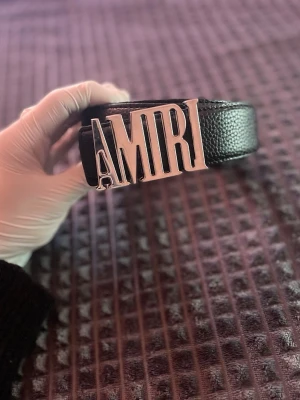 Svart läderbälte från Amiri - Säljer ett svart bälte i äkta läder från Amiri med en stor, silverfärgad metallspänne där det står AMIRI. Bältet har en stilren och modern look med textur i lädret och passar perfekt till jeans eller byxor.