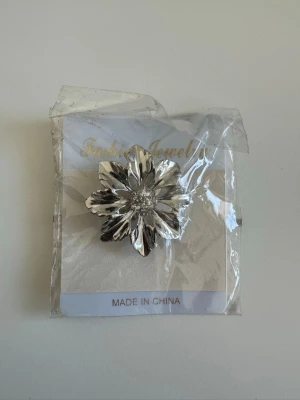 Silverfärgad blomma brosch - Superfin brosch i form av en blomma i blankt silvrigt material med glittriga stenar i mitten. Perfekt att sätta på blusen/kavajen/jackan/klänningen för att ge en extra touch. Helt NY i förpackning. 