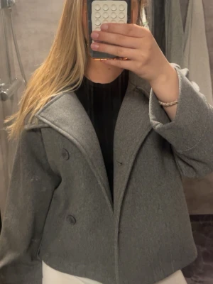 Grå kappa - Stilren grå kappa i kort modell med bred krage och dubbelknäppning framtill. Kappan har en rak passform och är tillverkad i ett mjukt tyg som ger en klassisk look. Använd ett par fåtal gånger 💞