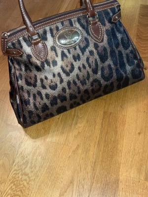 Roberto Cavalli Leopardmönstrad handväska (nyskick)  - Snygg handväska från Roberto Cavalli i leopardmönstrat tyg med bruna läderdetaljer och handtag. Väskan har en metallbricka med logga framtill och dragkedja upptill. Perfekt för dig som vill sticka ut med ett djurmönstrat statement. 100% äkta, ny skick.