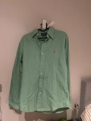 Grön rutig skjorta från Ralph Lauren - Snygg grön och vit rutig skjorta från Ralph Lauren med klassisk krage och custom fit-passform. Skjortan har långa ärmar, knappar framtill och den ikoniska broderade loggan på bröstet. Perfekt för en clean och stilren look.