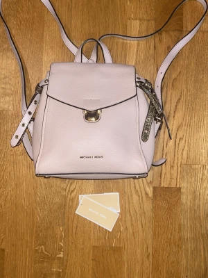 Ljusrosa ryggsäck Michael Kors (ny skick) - Snygg ljusrosa ryggsäck från Michael Kors i strukturerat läder. Väskan har guldfärgade metalldetaljer, justerbara axelband och ett praktiskt handtag upptill. Stängs med lock och spänne framtill. Perfekt för dig som vill ha en stilren och trendig accessoar. 100% äkta samt ny skick.