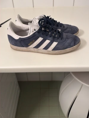Adidas Gazelles - Snygga Adidas Gazelle sneakers i blå mocka med vita detaljer och klassiska tre ränder på sidan. Låg modell med vit sula och guldfärgad text på sidan. Perfekta för en avslappnad och trendig stil.