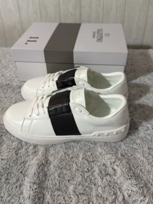Valentino Garavani vita sneakers 43 - Snygga vita sneakers från Valentino Garavani med bred svart läderdetalj över vristen. Skorna har vita snören, gummisula med mönster och subtila nitar på hälen. Märkeslogga syns på plösen och innersulan. Perfekta för en trendig och stilren look.