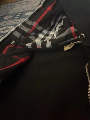 Svart hoodie med rutigt foder - Svart hoodie med dragkedja och stor huva. Insidan av huvan och öppningen har ett rutigt foder i svart, vitt och rött. Hoodien har långa ärmar och är tillverkad i ett mjukt material, perfekt för en avslappnad look.