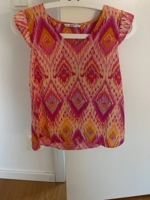 Färgglad mönstrad topp från ONLY - Säljer en färgglad topp från ONLY med geometriskt mönster i rosa, lila, orange och beige. Toppen har korta ärmar och en luftig passform, perfekt för varma dagar. Materialet känns lätt och bekvämt mot huden.
