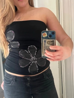 Svart tubtopp med blommor - Säljer en svart tubtopp med stora blommor i silver och glitterdetaljer. 