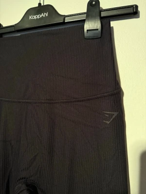 Svarta tights från Gymshark - Säljer ett par svarta tights från Gymshark med hög midja och diskret logga på höften. Tillverkade i stretchigt, ribbat material som sitter skönt och följer kroppen perfekt. Perfekta för gymmet eller annan träning. Byxorna är köpta för 549kr och aldrig använda 