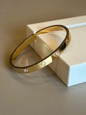 Guldfärgat armband med stenar - Snyggt guldfärgat armband i metall med infattade klara stenar och ingraverade blommönster. Armbandet har en stilren, rundad form och passar perfekt för dig som gillar eleganta accessoarer med lite extra bling.