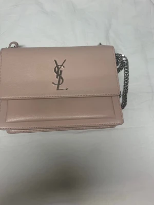 Ljusrosa Saint Laurent handväska - Säljer en ljusrosa handväska från Saint Laurent med silverfärgad kedja och ikoniskt YSL-logo på framsidan. Väskan är tillverkad i slätt läder och har ett stilrent, rektangulärt format. Perfekt för dig som gillar exklusiva accessoarer.//ingen kvitto och inga paket min ex man han köpte för 15 år sedan