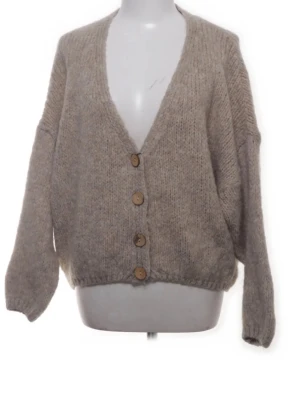 Beige stickad kofta med knappar - Mysig beige stickad kofta med djup v-ringning och fyra stora träknappar framtill. Koftan är tillverkad av en blandning av akryl, polyamid, mohair och ull, vilket gör den både mjuk och varm. Perfekt för lager-på-lager och chill dagar.