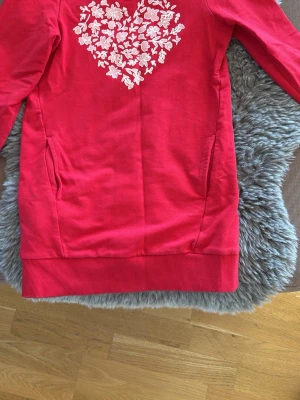 Rosa hoodie med vit hjärttryck - Cool hoodie i stark rosa färg med ett stort vitt hjärttryck av blommor på bröstet. Tröjan har ribbade muddar och två sidofickor. Perfekt för dig som gillar att sticka ut och vill ha något unikt i garderoben.