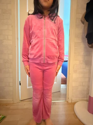 Rosa hoddie set från Lindex  - Mysigt loungewear-set i rosa velour med zip-tröja och matchande byxor. Tröjan har huva, dragkedja och fickor framtill. Byxorna är raka och mjuka, perfekta för chill hemma. Materialet är lent och stretchigt för extra komfort.