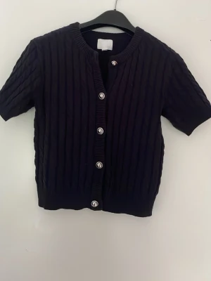 Svart kabelstickad kofta från H&M - Kortärmad svart kabelstickad kofta från H&M med silvriga knappar framtill. Koftan har rund halsringning och ribbade kanter. Perfekt att slänga över en t-shirt eller linne för en snygg lager-på-lager-look.