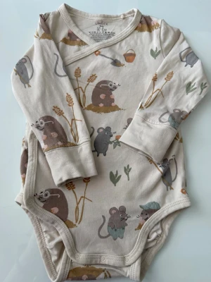 Söt body med musmotiv från Lindex - Beige långärmad babybody från Lindex med gulligt tryck av möss, växter och små detaljer i brunt, grått och grönt. Omlottknäppning och tryckknappar i grenen. Tillverkad i mjuk bomull med lite elastan för extra komfort.
