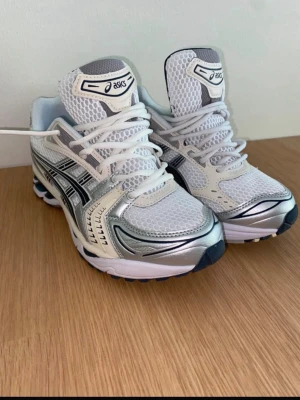  Asics Kayano 14 - Snygga sneakers från Asics i vitt och silver med svarta detaljer. Ovandelen är i mesh och syntetmaterial för bra andningsförmåga och komfort. Klassisk Asics-logga på sidan och tungspetsen. Perfekta för dig som gillar sportig och trendig stil.