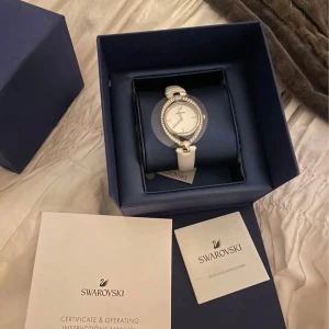 Vit klocka från Swarovski - Vit klocka från Swarovski med stenar runt urtavlan. Klockan har ett armband i vitt och kommer i förpackning. Aldrig använd så helt ny. Kan behöva bytas batteri.
