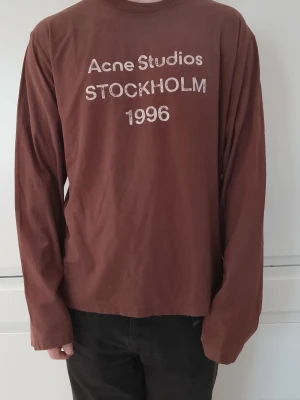 Brun långärmad topp Acne Studios - Säljer en brun långärmad topp från Acne Studios med tryck på bröstet där det står 'Acne Studios STOCKHOLM 1996'. Skön passform och tillverkad i mjuk bomull. Perfekt för dig som gillar minimalistisk och stilren design.