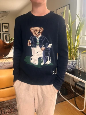 Mörkblå stickad tröja Polo Bear RL - Säljer en mörkblå stickad tröja från Ralph Lauren med den ikoniska Polo Bear på bröstet. Tröjan har rund halsringning, ribbade muddar och RL-broderi nedtill. Perfekt för dig som gillar klassisk preppy stil och vill sticka ut med en unik detalj.